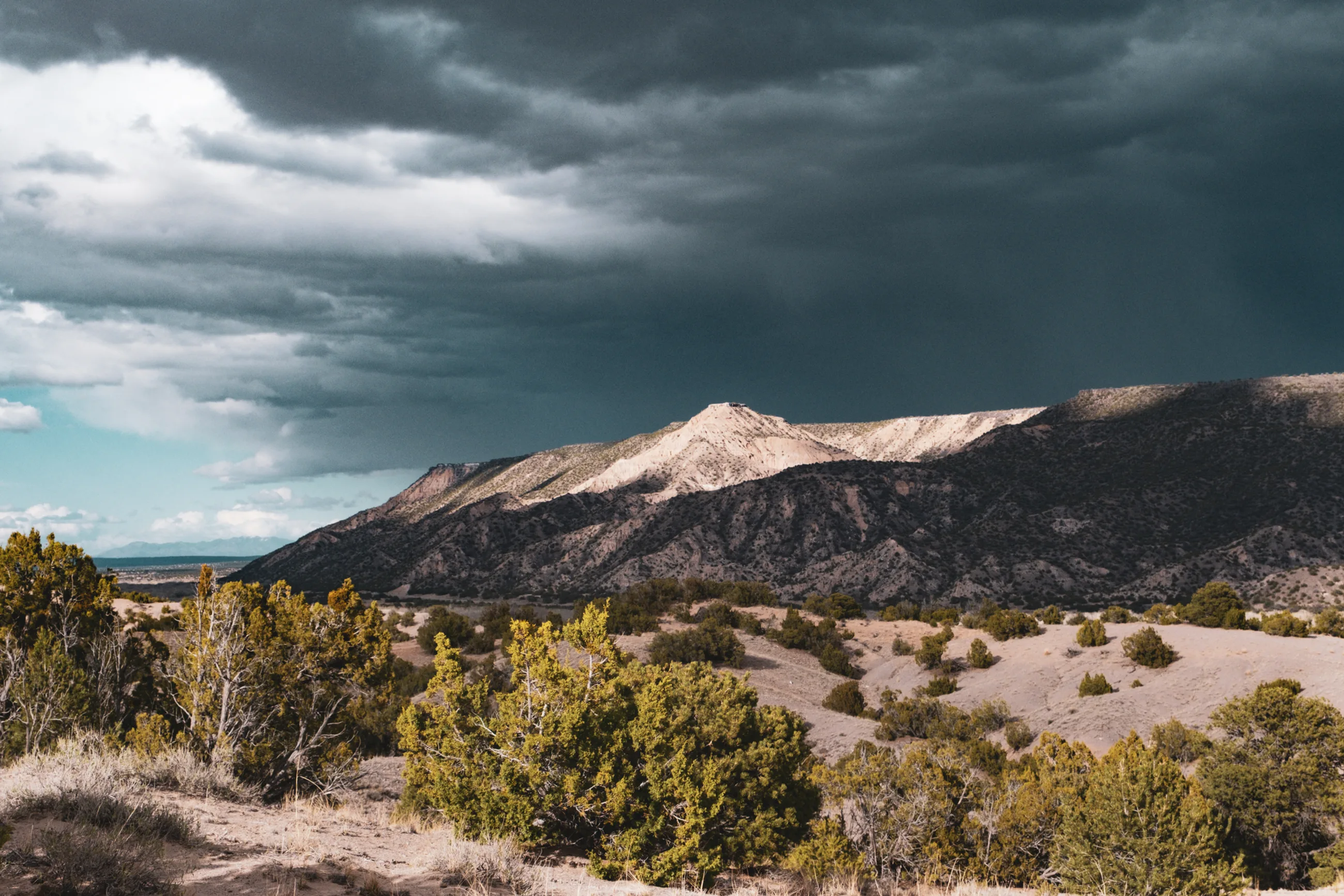 Storm clouds gathering over a pale mesa and juniper-dotted hills in the New Mexico high desert